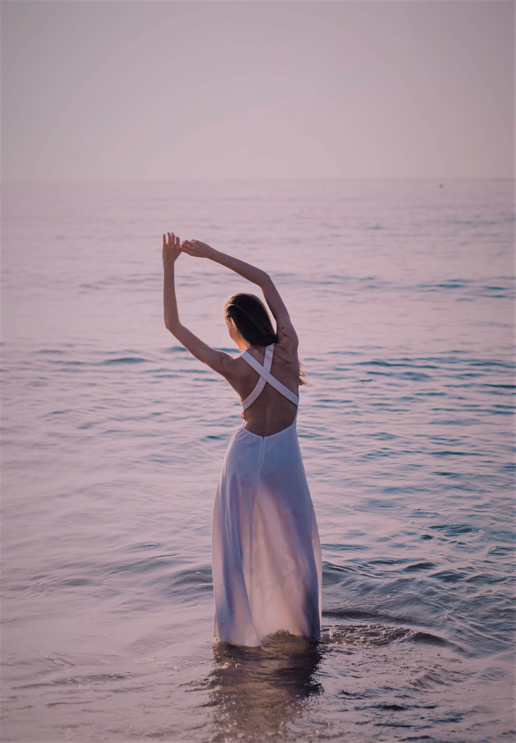 Dies ist ein foto, das eine Frau zeigt, die mit dem Rücken zur Kamera im Meer steht. Sie hat ihre Arme über den Kopf genommen und neigt sich leicht zur Seite.