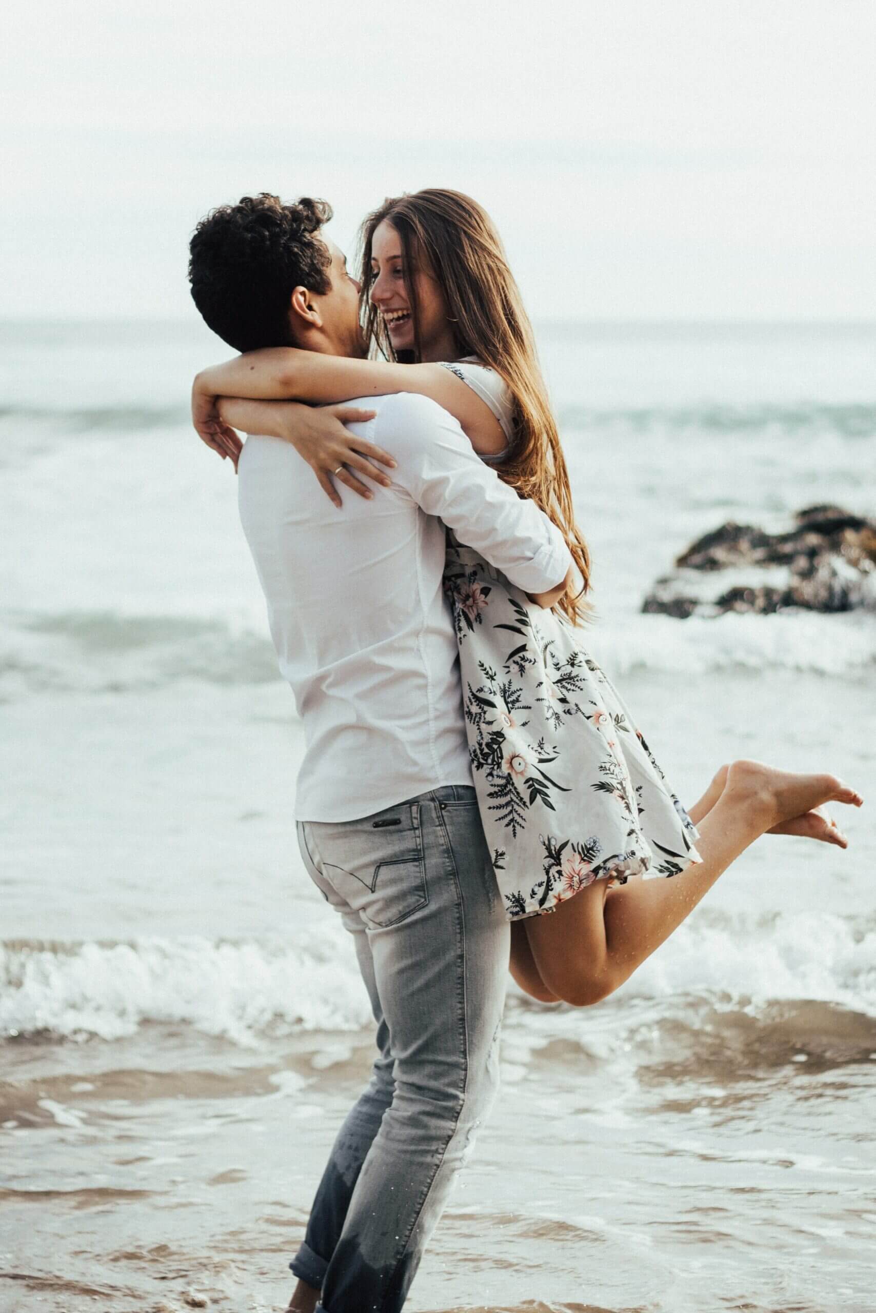 Dies ist ein Foto, das einen Mann und eine Frau am Meer zeigt. Der Mann hebt die Frau hoch, beide lächeln und umarmen sich.