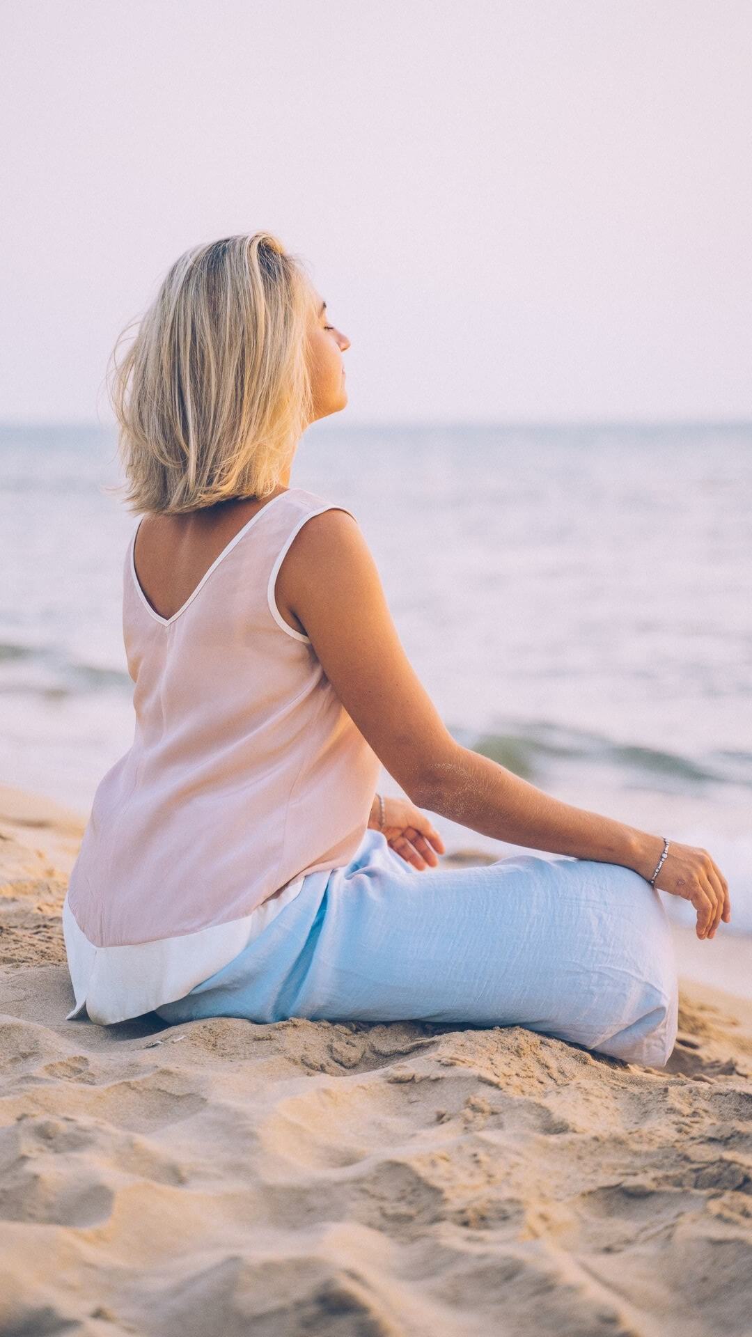 Dies ist ein Foto, das eine Frau zeigt, die im Sand sitzt und meditiert. Sie hat halblange blonde Haare und hat die Augen geschlossen. Sie ist dem Wasser zugewandt, trägt ein rosa Oberteil und eine helle Hose.