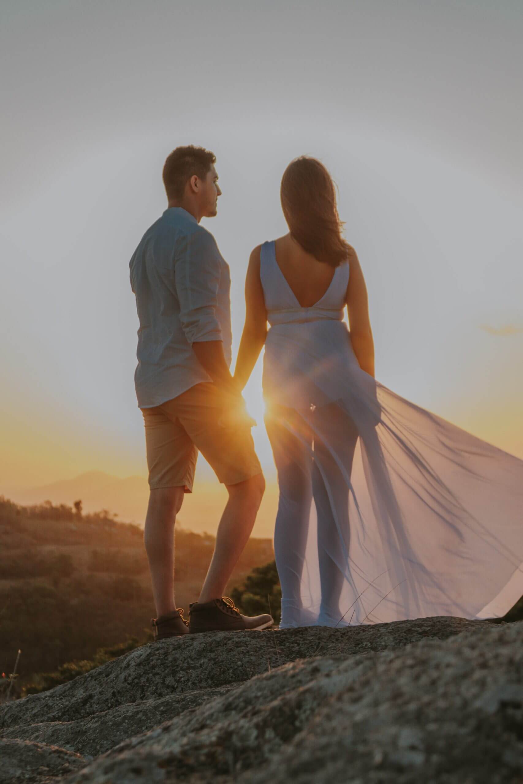 Auf diesem Bild sieht man einen Mann und eine Frau, die mit dem Rücken zur Kamera stehen. Sie stehen auf einem Hügel, halten Händchen und schauen Richtung Sonnenuntergang.